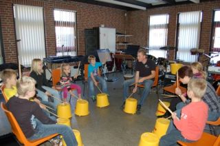 Kinderen met leerkracht in kring, gebruiken een emmer en houten stokken als drums